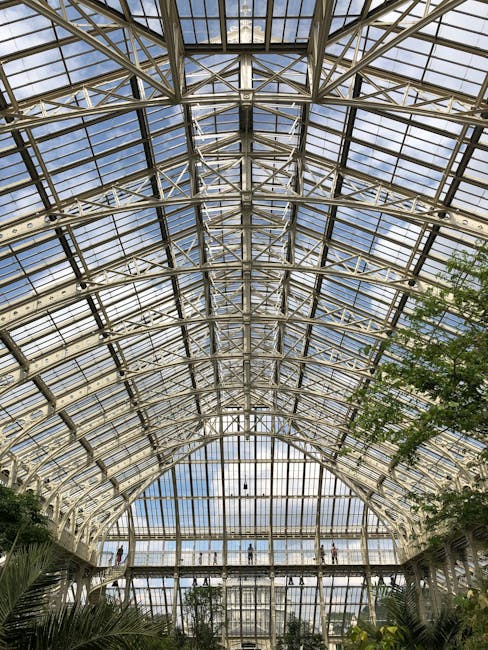 Inside a large glass greenhouse with an intricate metal framework supporting curved panels of glass, allowing natural light to flood the space. The structure features multiple levels and walkways wher